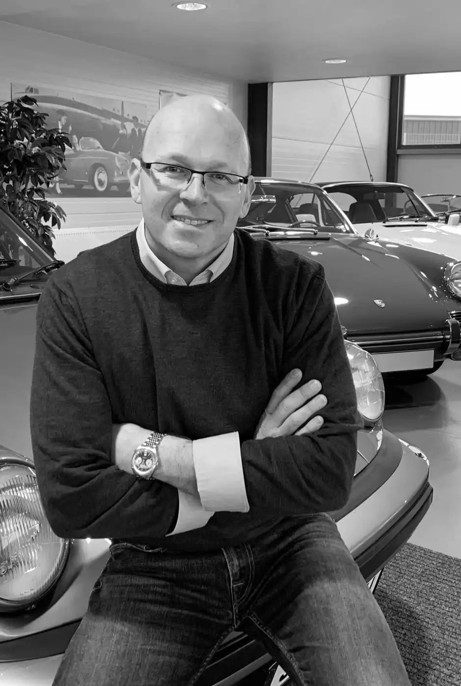 Ein Portrait von Dennis Nachtigal auf einer Motorhaube sitzend inmitten seiner Fahrzeughalle